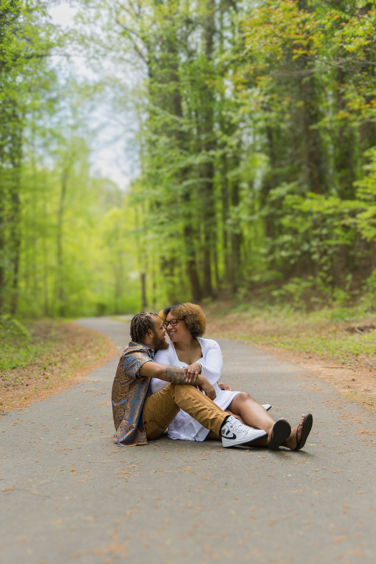 Two figures sit intimately together on a scenic forest path surrounded by lush green trees on a bright day.