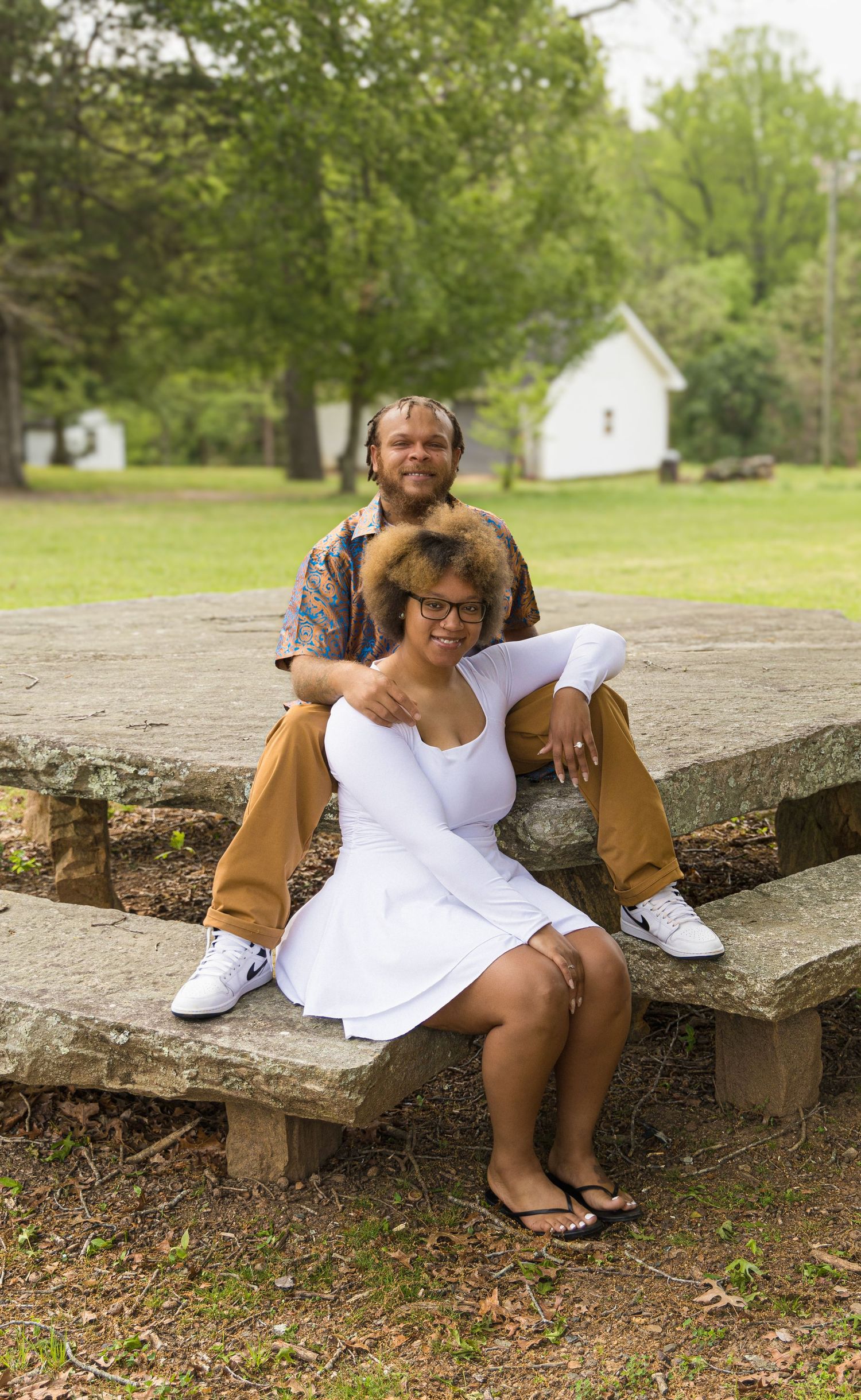 A couple relaxes together on rustic wooden steps outdoors with a white building and green trees in the background.