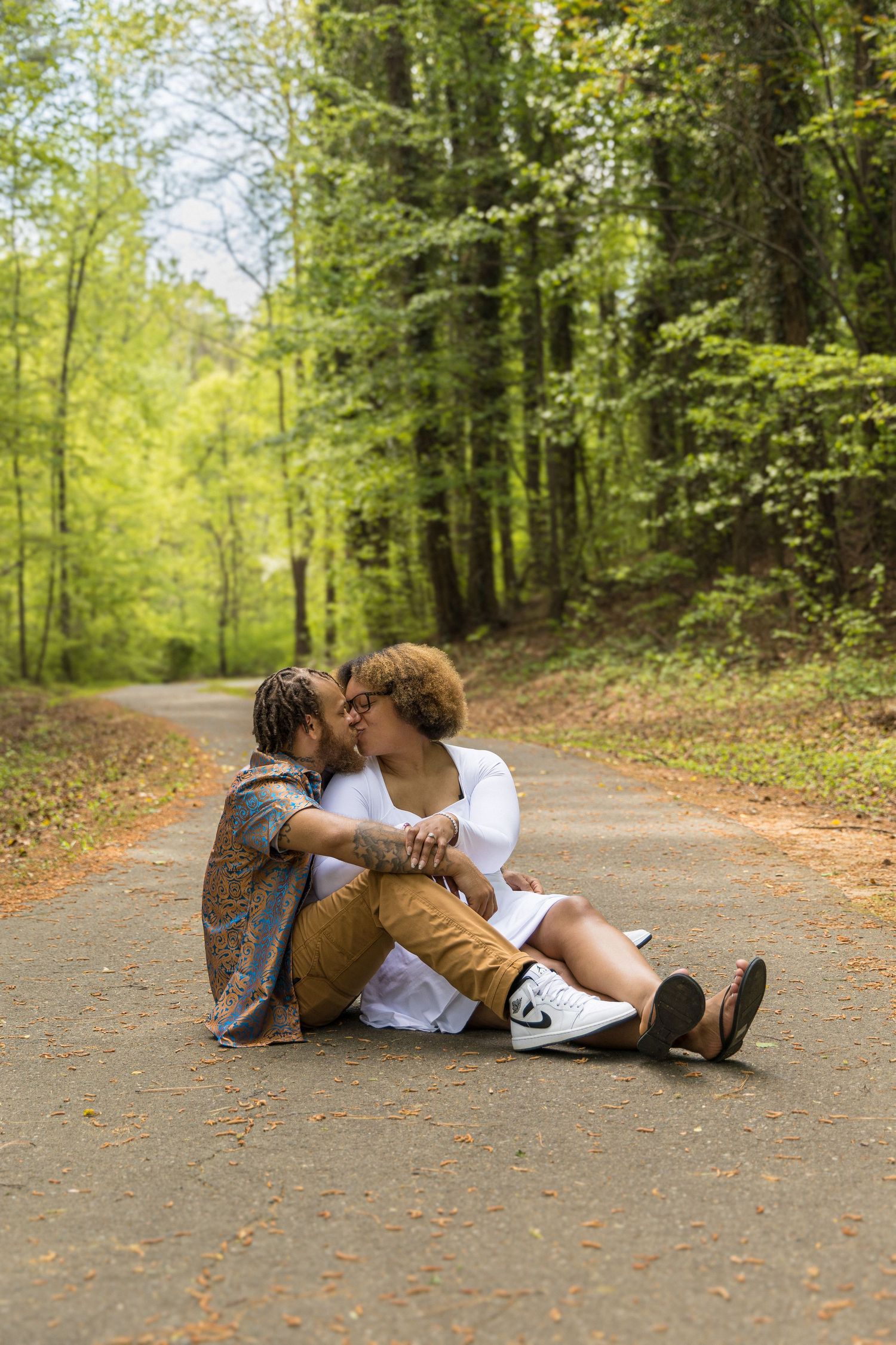 Two people share an intimate moment sitting together on a forest path surrounded by lush green trees on a sunny day.