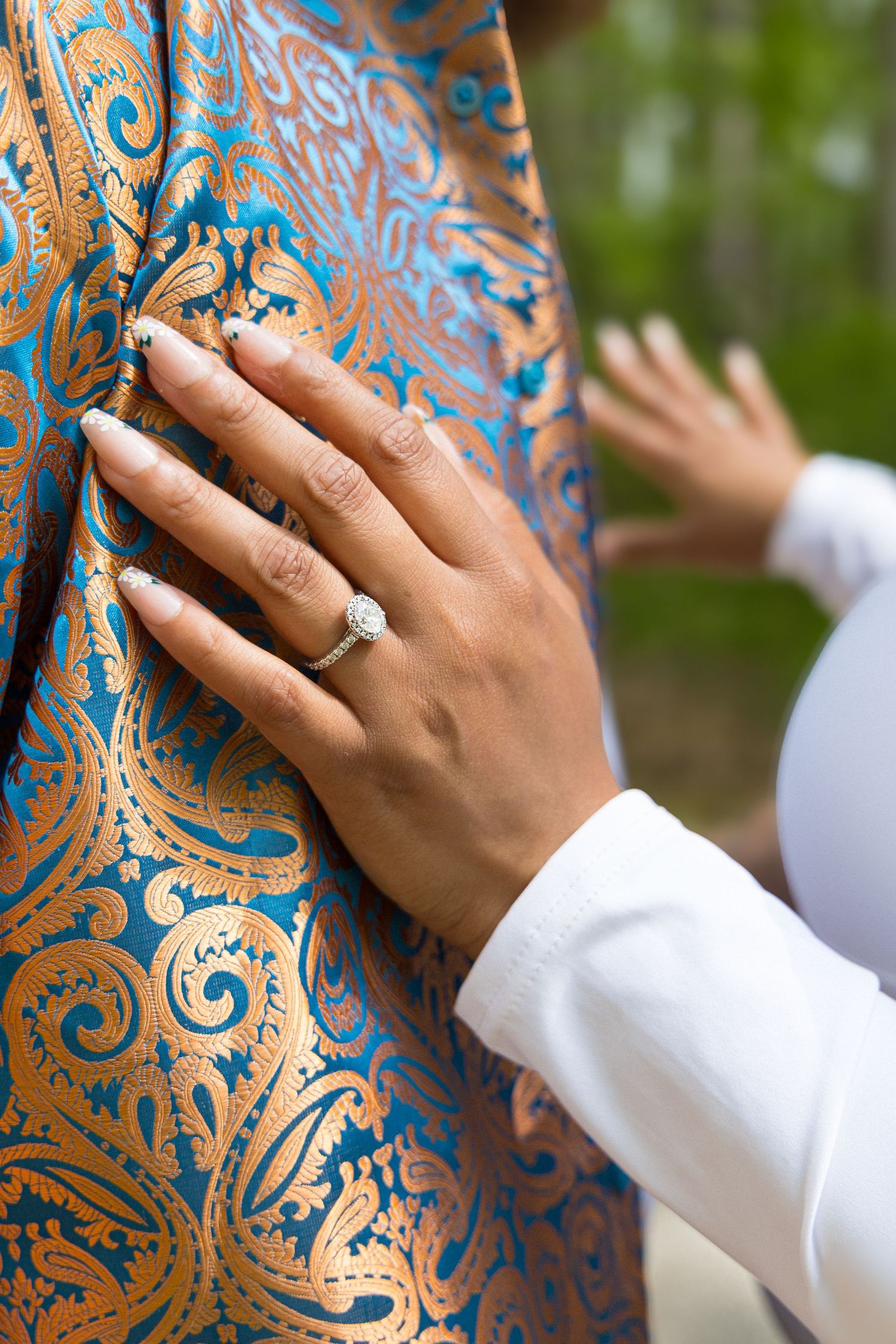 Hand wearing white sleeve displays engagement ring against ornate blue and gold patterned fabric backdrop.