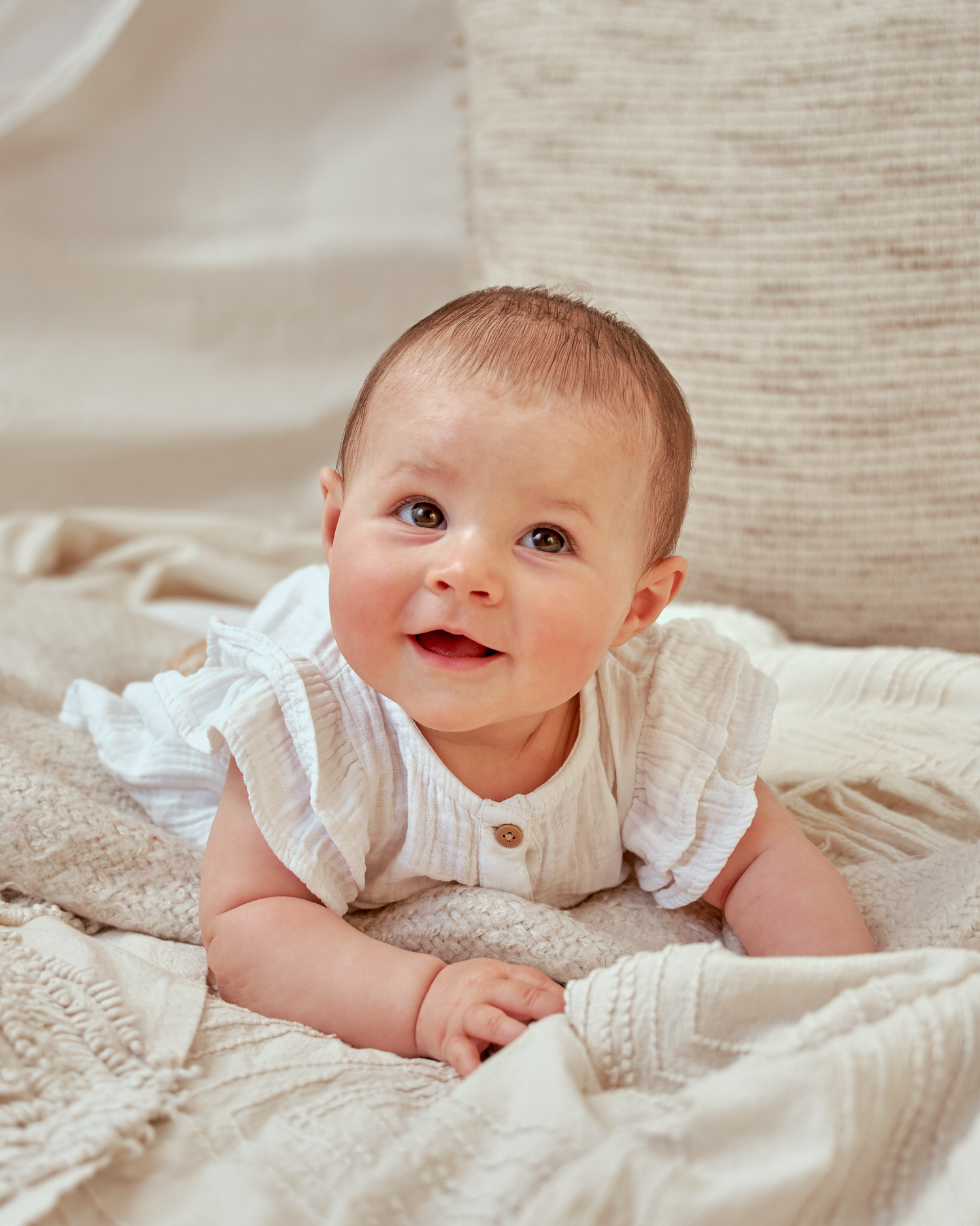 Smiling baby in white ruffled outfit lying on textured beige blanket.