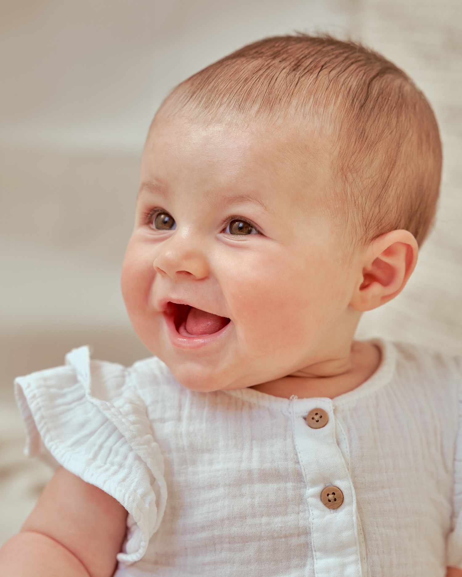 Baby wearing white ruffled outfit smiles brightly at the camera.