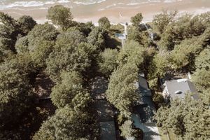 Aerial view of a coastal house surrounded by lush green trees with sandy beach and ocean waves visible.