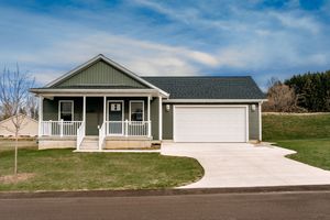 A small green ranch-style house with a covered front porch and attached two-car garage sits on a grassy lot.