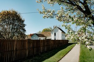 White house with wooden fence along a sidewalk path on a sunny spring day with blooming cherry blossoms overhead.