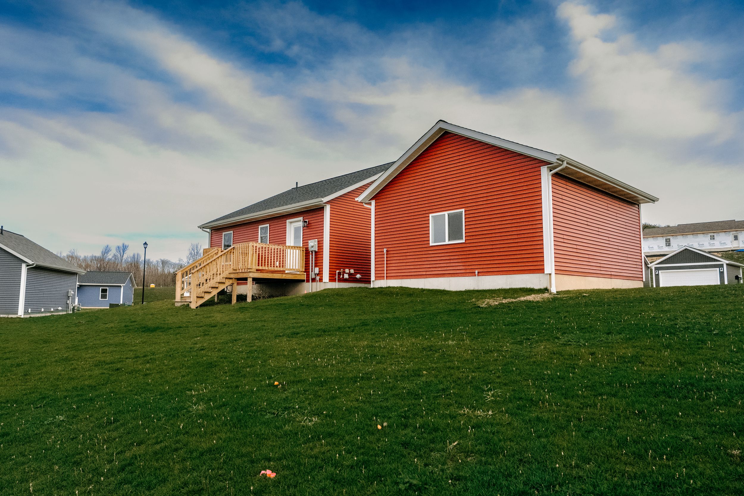 Red ranch house with covered porch sits on green hillside under dramatic blue sky with wispy clouds.