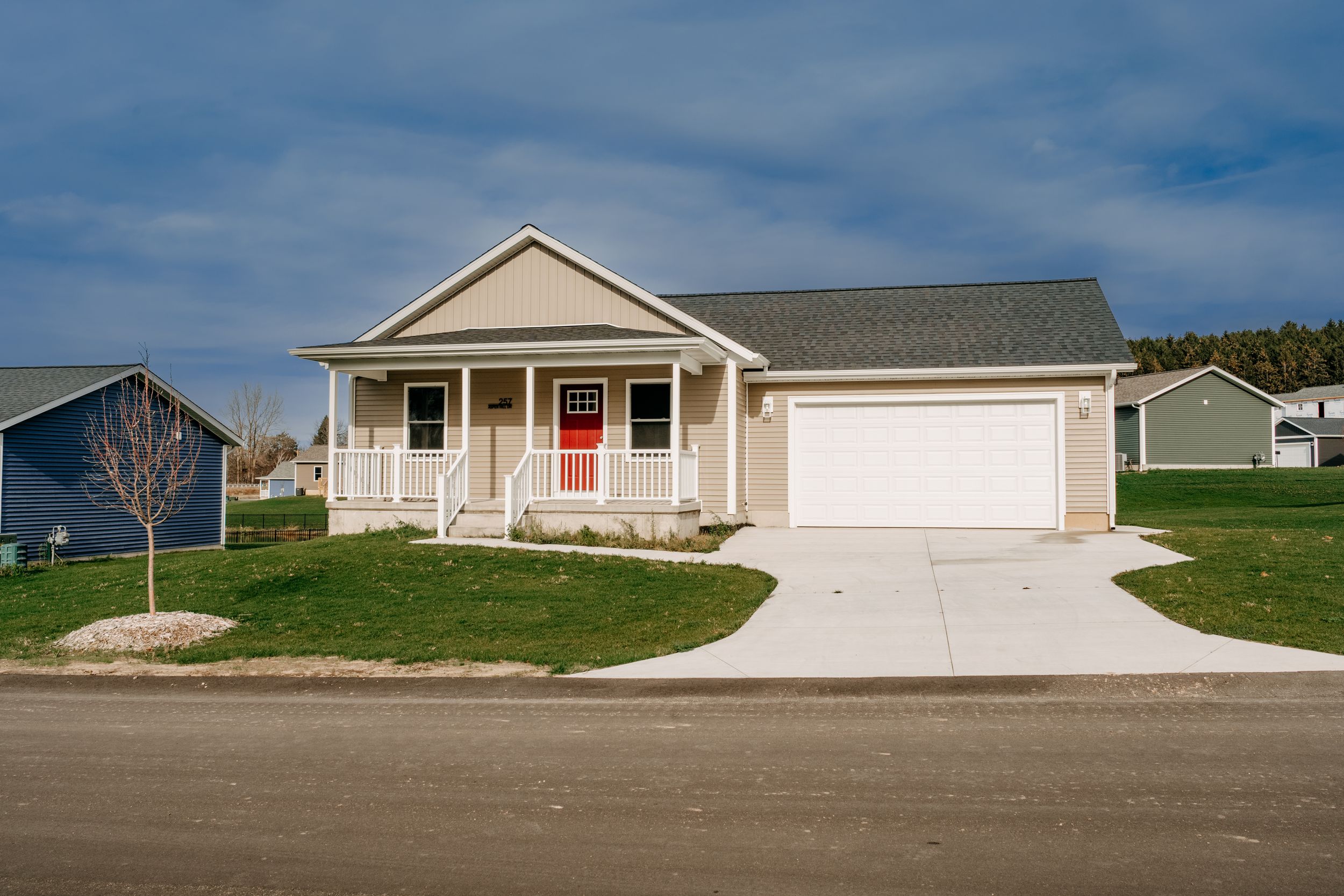 Small beige ranch style home with red door, white trim, and attached two car garage on landscaped suburban lot.