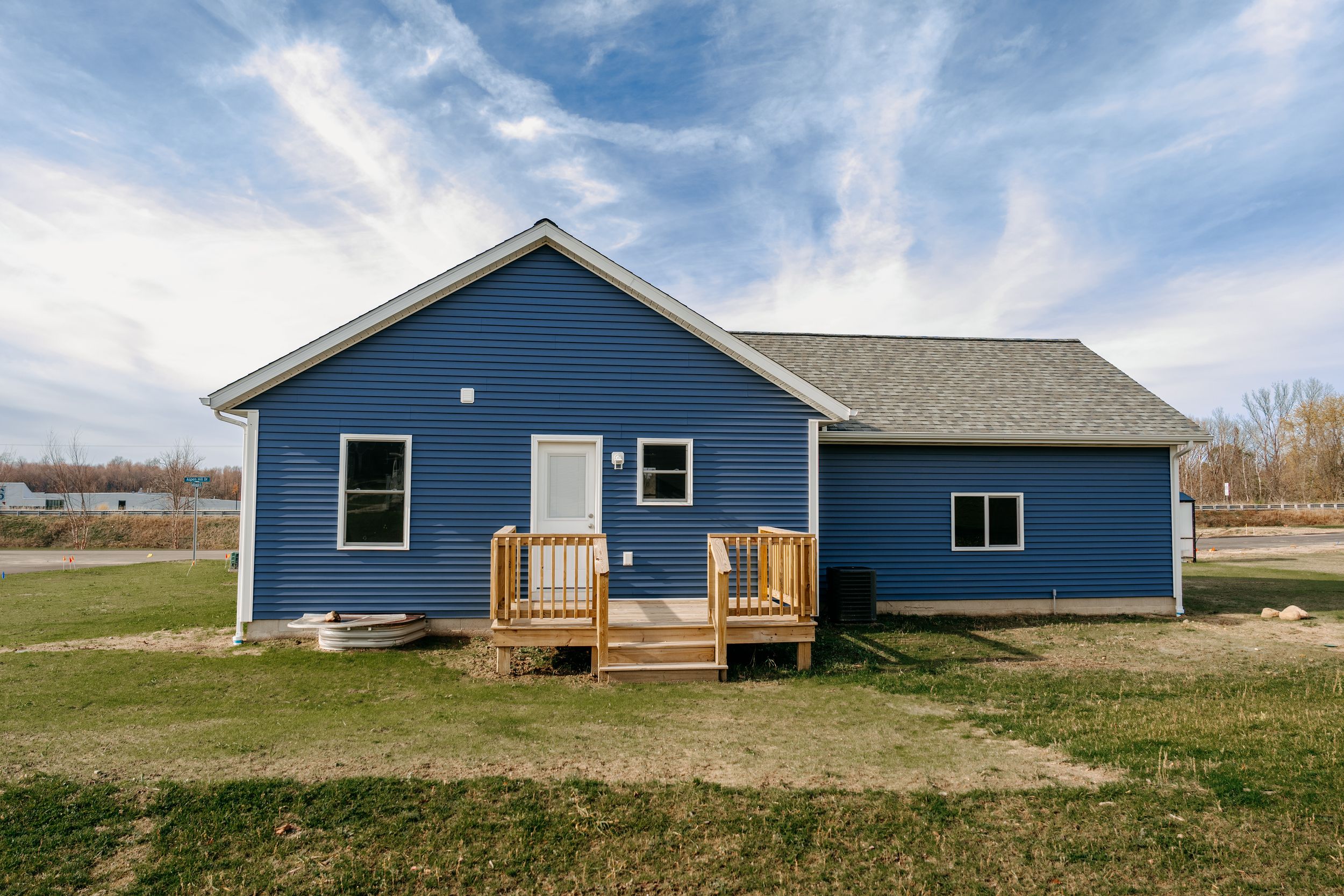 A modern blue ranch-style house with a wooden deck and black trim sits on a grassy lot under blue skies.