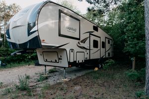 A large fifth-wheel RV camper trailer parked in a wooded campsite with trees in the background.