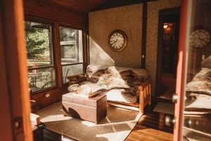 Sunlit wooden bedroom interior with vintage clock and luggage showing warm morning light streaming through windows.
