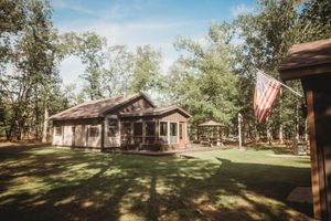Rustic wooden cabin with American flag surrounded by tall trees on a sunny day in a serene forest setting.