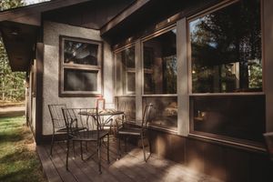 A cozy rustic cabin porch with metal chairs and sunlight casting shadows through screened windows.
