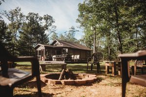 Rustic wooden cabin in the woods with a covered porch and outdoor fire pit area surrounded by tall trees.