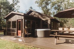 A rustic wooden cabin with a screened porch and hot tub on the deck surrounded by trees in natural setting.
