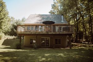 Rustic two-story house with large wooden deck surrounded by trees in a wooded backyard setting.