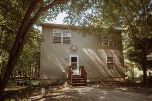 A tan two-story home with wooden steps sits nestled among leafy trees casting dappled shadows on its exterior.