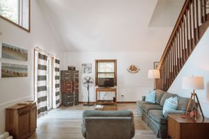 A cozy living room interior with a teal sofa, wooden staircase, high ceiling, and natural light from tall windows.