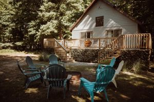 Rustic white cabin in the woods with turquoise Adirondack chairs arranged around a fire pit on a sunny afternoon.
