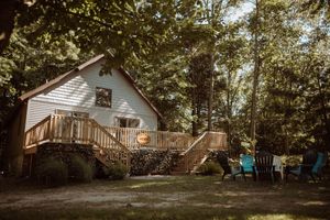 Cozy white cabin in the woods with a wooden deck and blue Adirondack chairs in the front yard.
