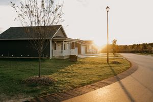 A cozy suburban house with white siding sits along a curved road at sunset with a streetlamp casting warm light.