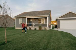 A cozy ranch-style home with gray siding, white trim, and a detached garage sits on a well-maintained green lawn at dusk.
