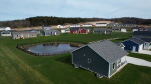 Aerial view of suburban development with houses surrounding a small pond and green lawns on a cloudy day.