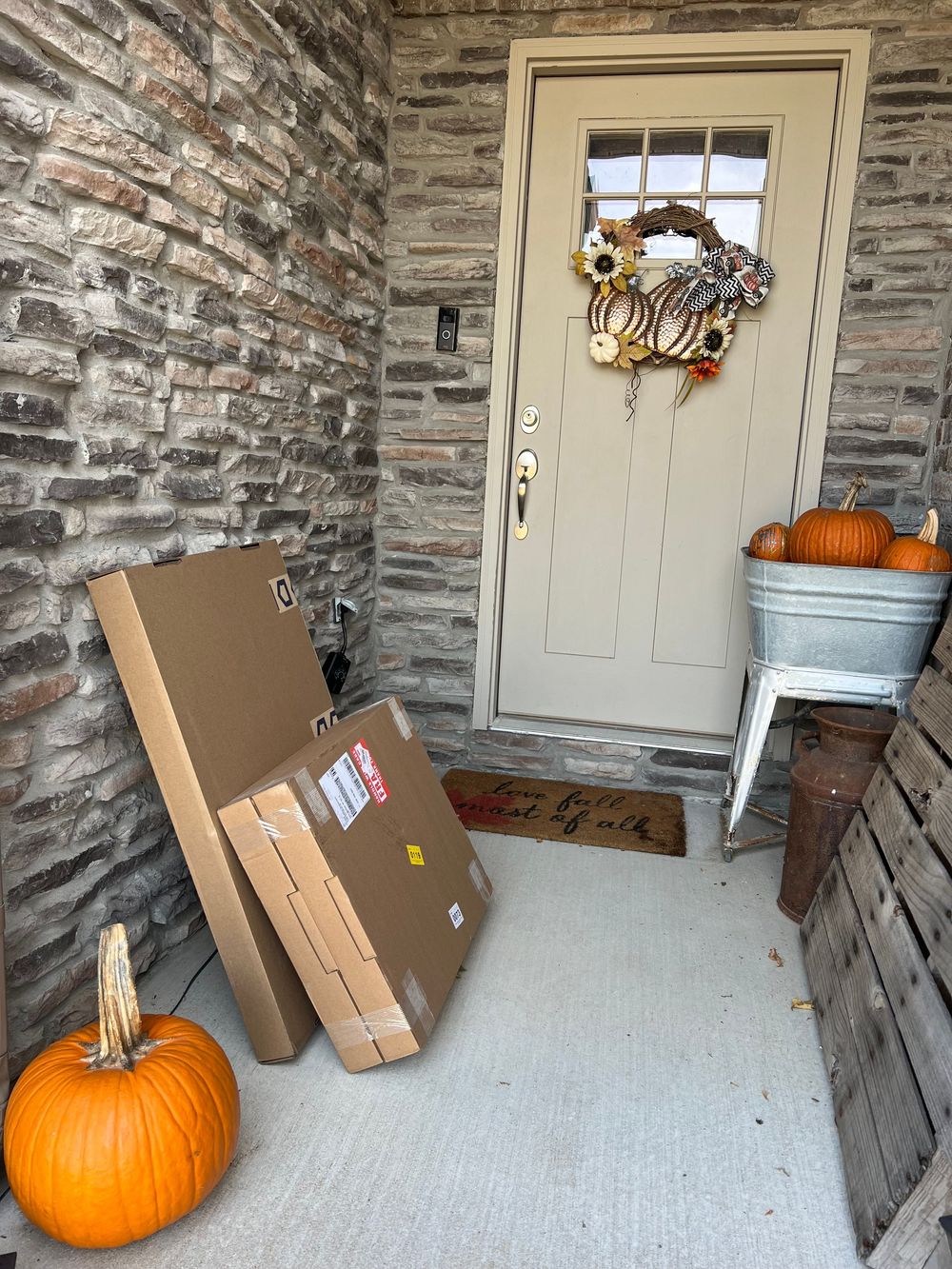 Series of cardboard boxes and a large orange pumpkin arranged on a stone-walled porch with white door.