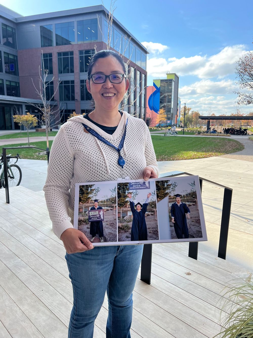 A person in a white mesh top and jeans holds up photos while standing on a concrete plaza on a sunny day.