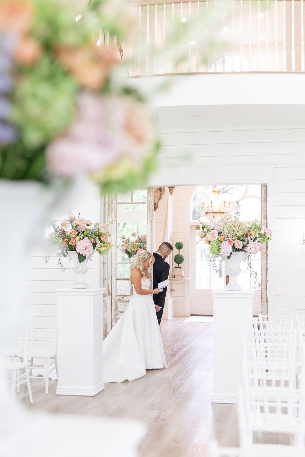 Bride and groom share an intimate moment at the entrance of a bright white wedding venue with floral arrangements.