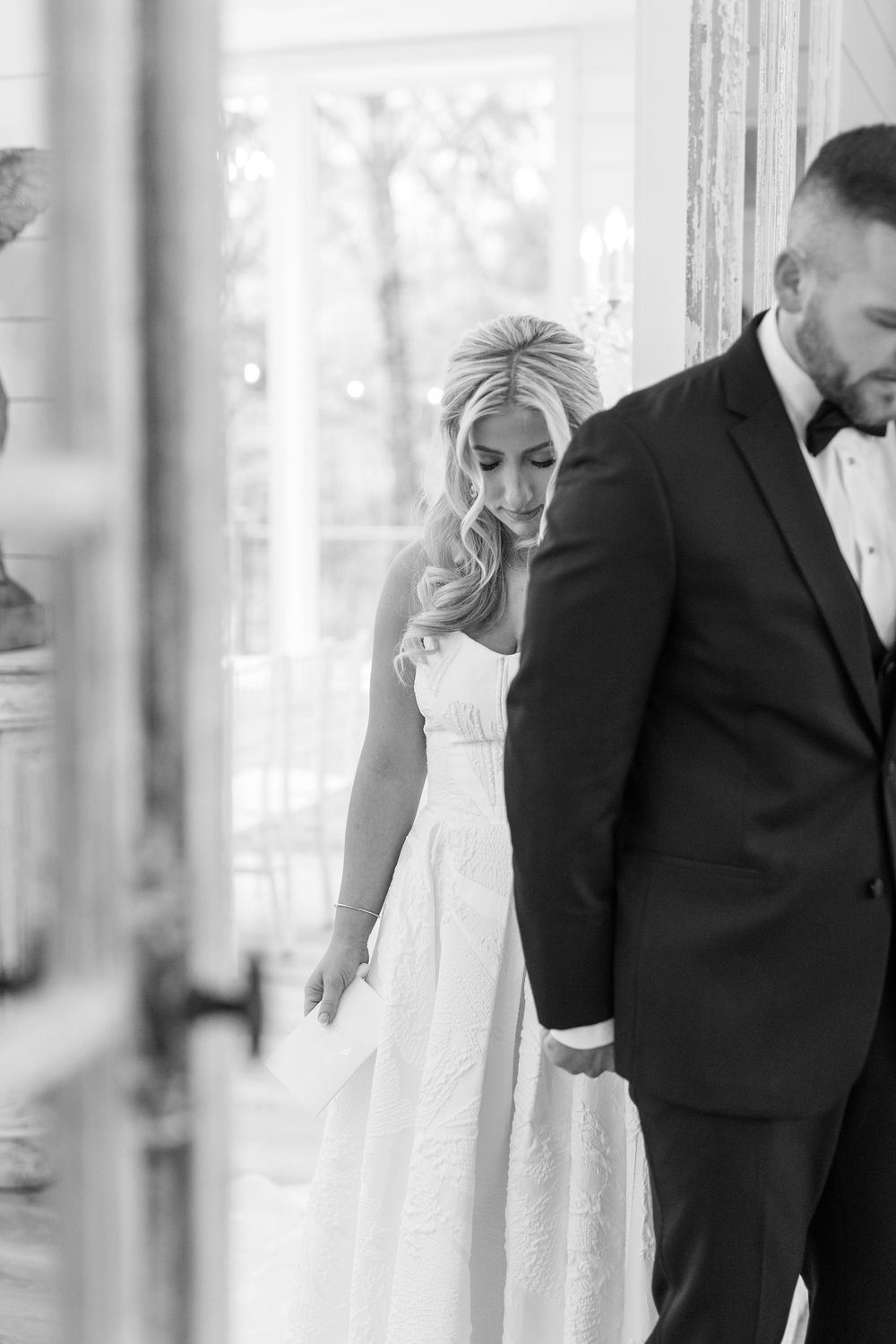 Black and white photo of bride in strapless gown and groom in tuxedo sharing quiet moment by window.