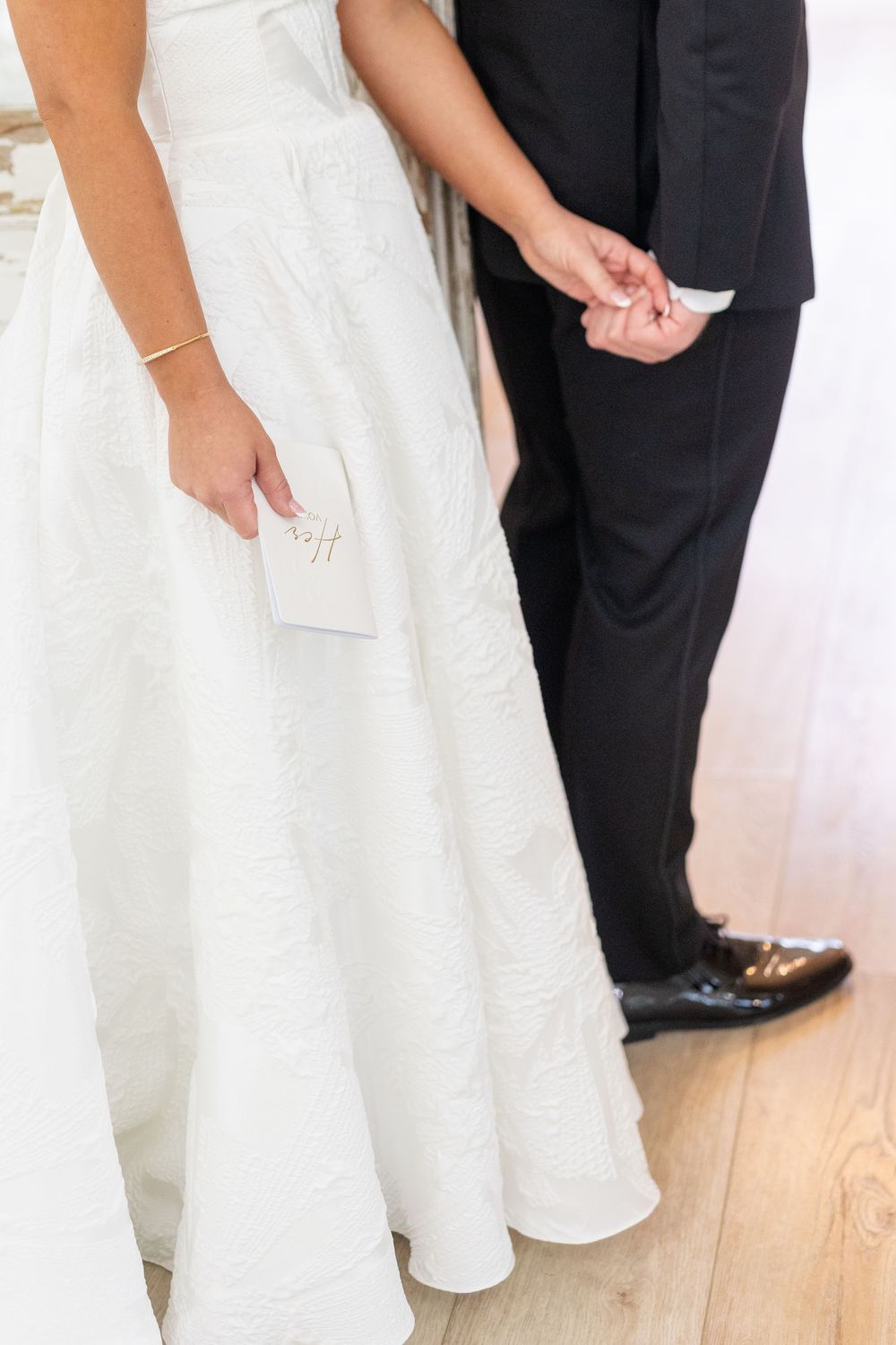 Close-up of bride in white gown holding vows booklet while groom in black suit holds her hand.