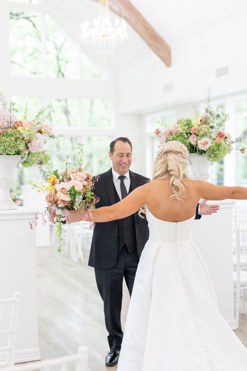 Groom smiles as bride opens arms for first look reveal in a bright white wedding venue with florals.