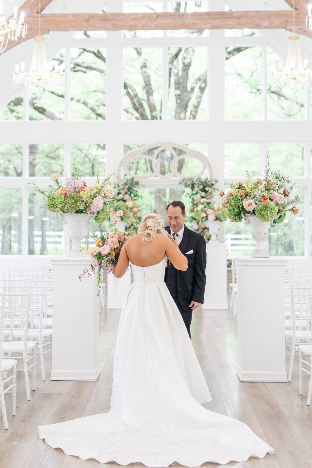 Bride in strapless gown with long train faces groom during first look in sunlit white wedding chapel.