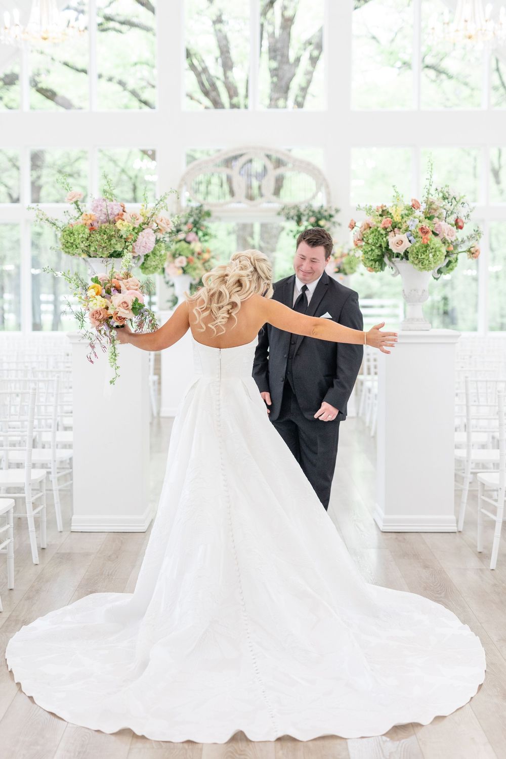Bride and groom share joyful first look moment in elegant white chapel with colorful floral arrangements.