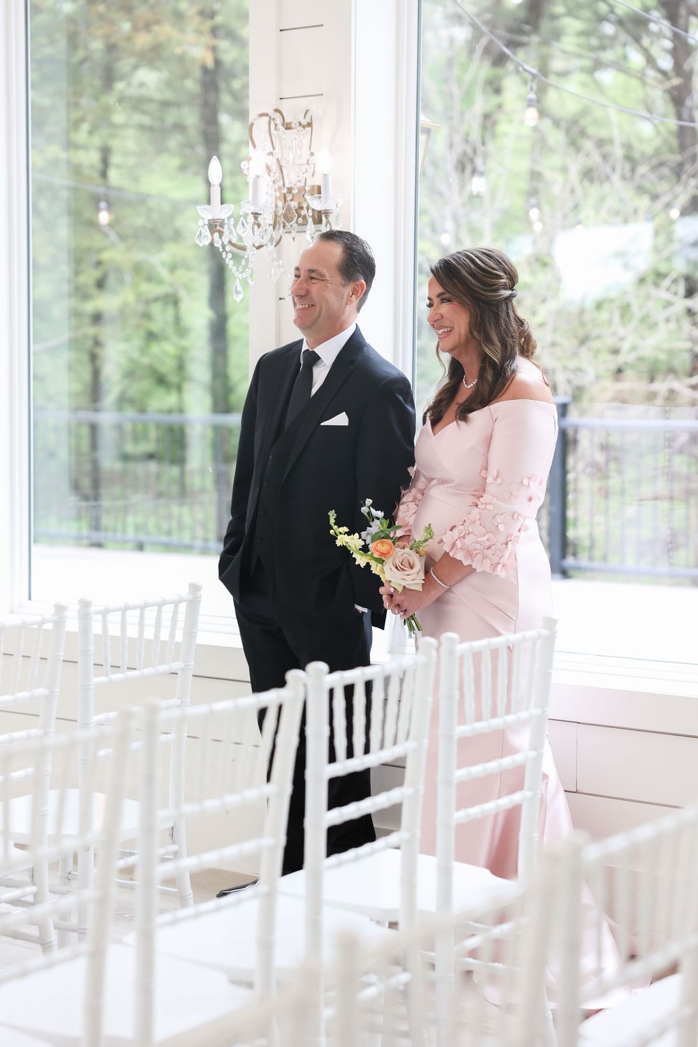 Smiling couple in formal attire stands among white Chiavari chairs in a bright windowed wedding venue.