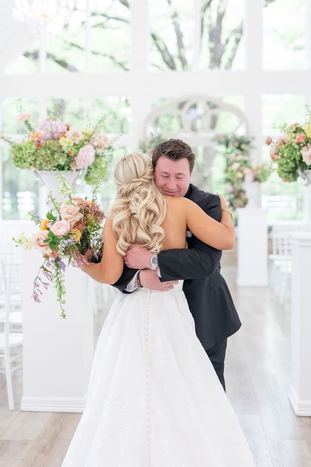 Emotional groom hugs bride in strapless gown during first look in a floral-decorated white wedding venue.