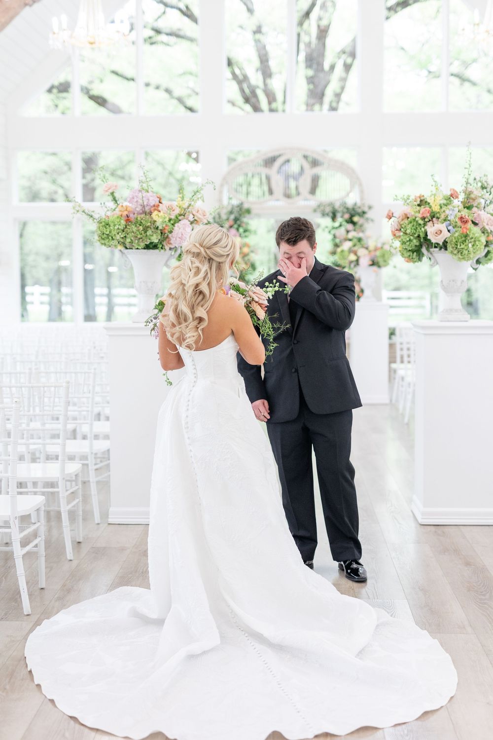 Tearful groom wipes eyes during emotional first look with bride in elegant white chapel with floral decor.
