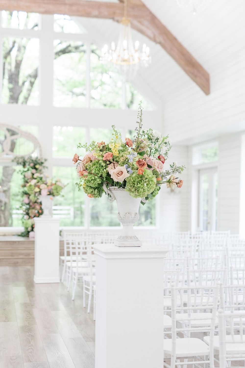 Elegant white wedding chapel with floral arrangements in urns lining the aisle and white chiavari chairs.
