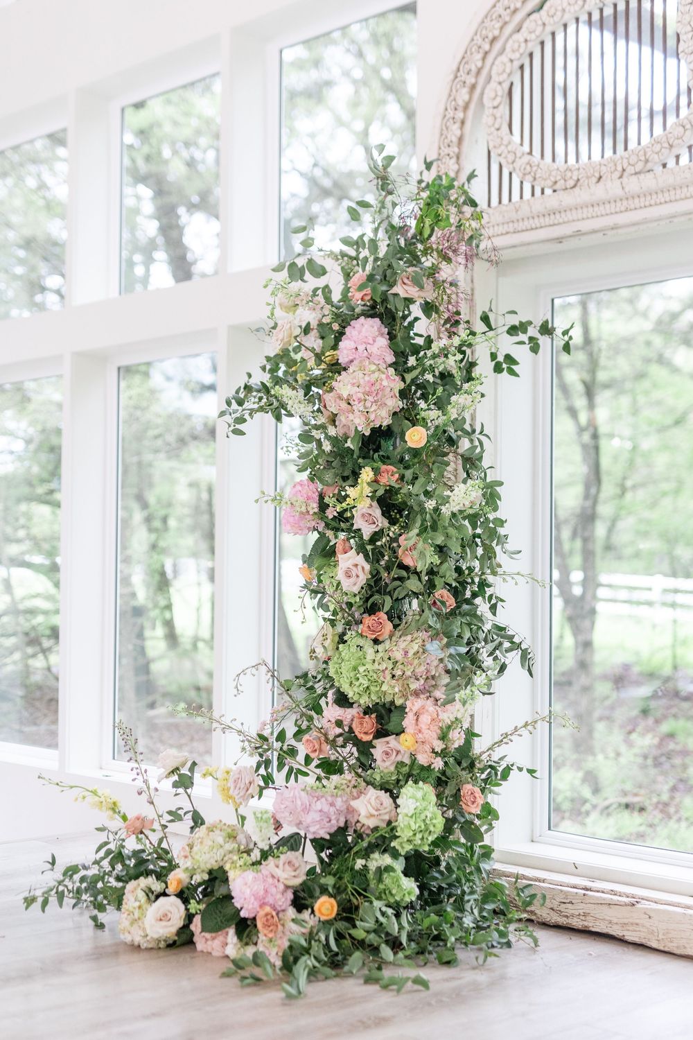 Lush floral column installation with pink hydrangeas, roses, and greenery beside an arched mirror.
