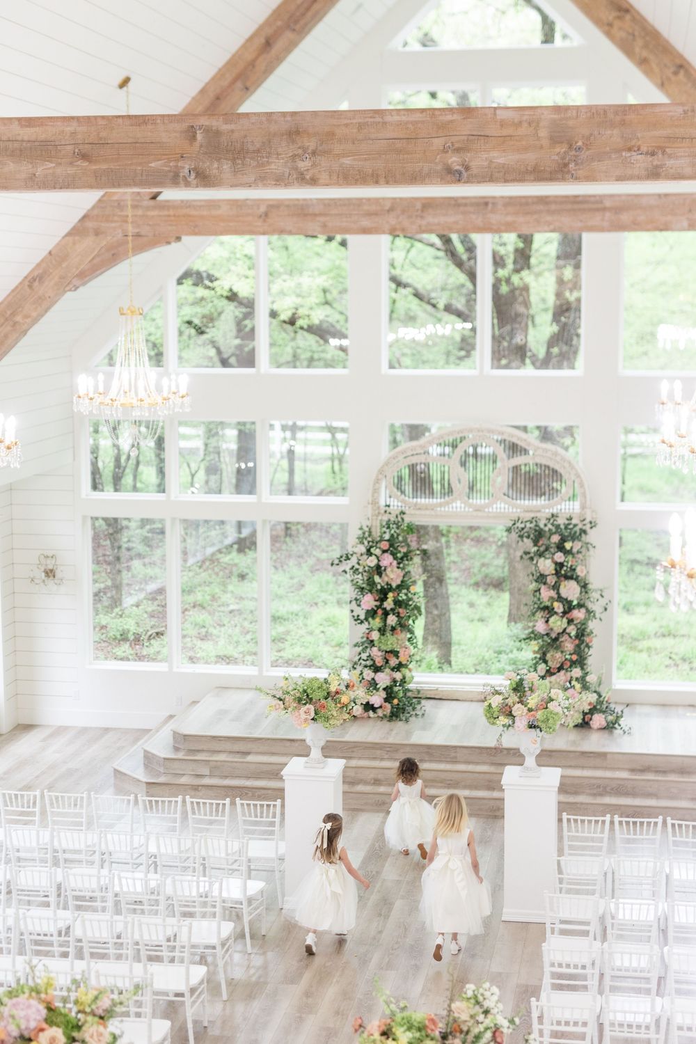 Bright white wedding venue with wood beam ceiling, chandeliers, and floral arch at the altar.