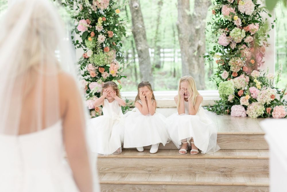 Three flower girls in white dresses react with surprise and joy upon seeing the bride.