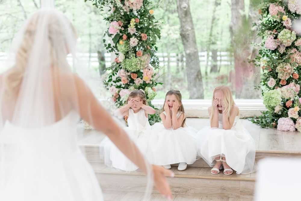 Flower girls seated on steps react with delight and awe as bride approaches in white gown.