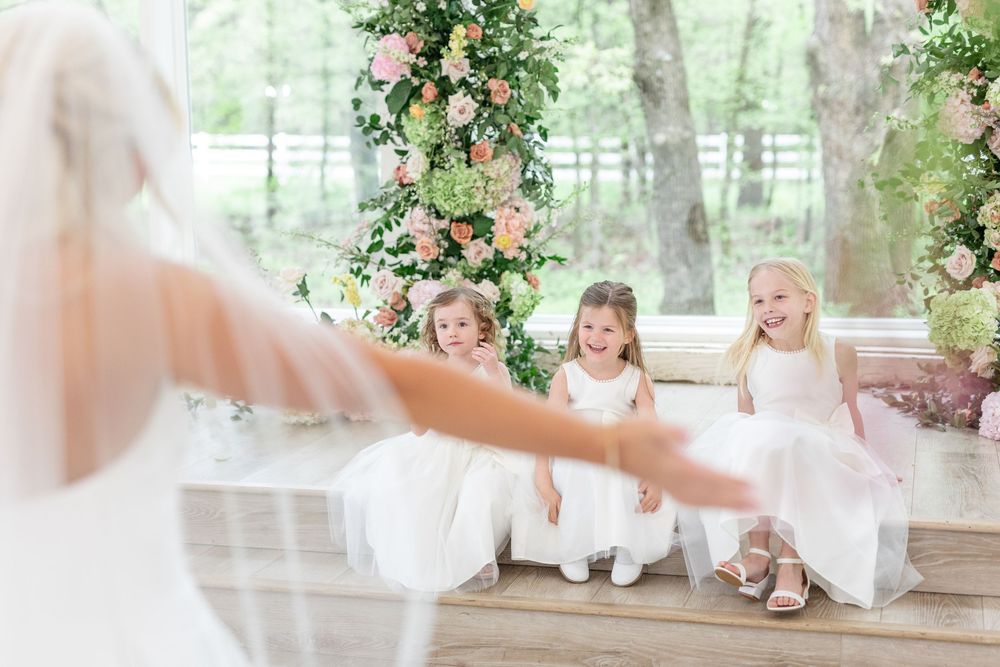 Three smiling flower girls in white dresses reach out excitedly toward the approaching bride.