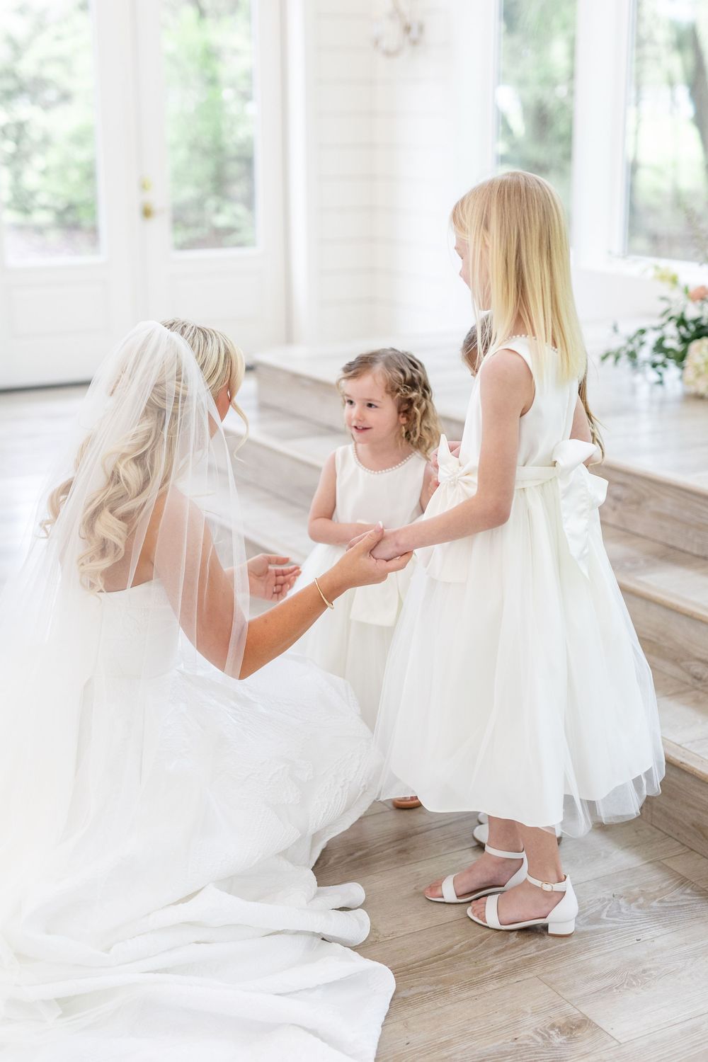 Bride in white gown kneeling to hold hands with two flower girls in a bright white venue.