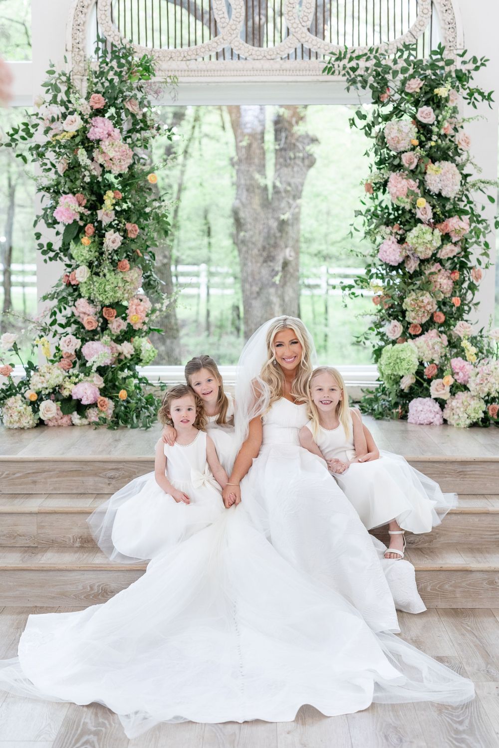 Bride in ballgown seated with three flower girls in front of lush floral arch ceremony backdrop.