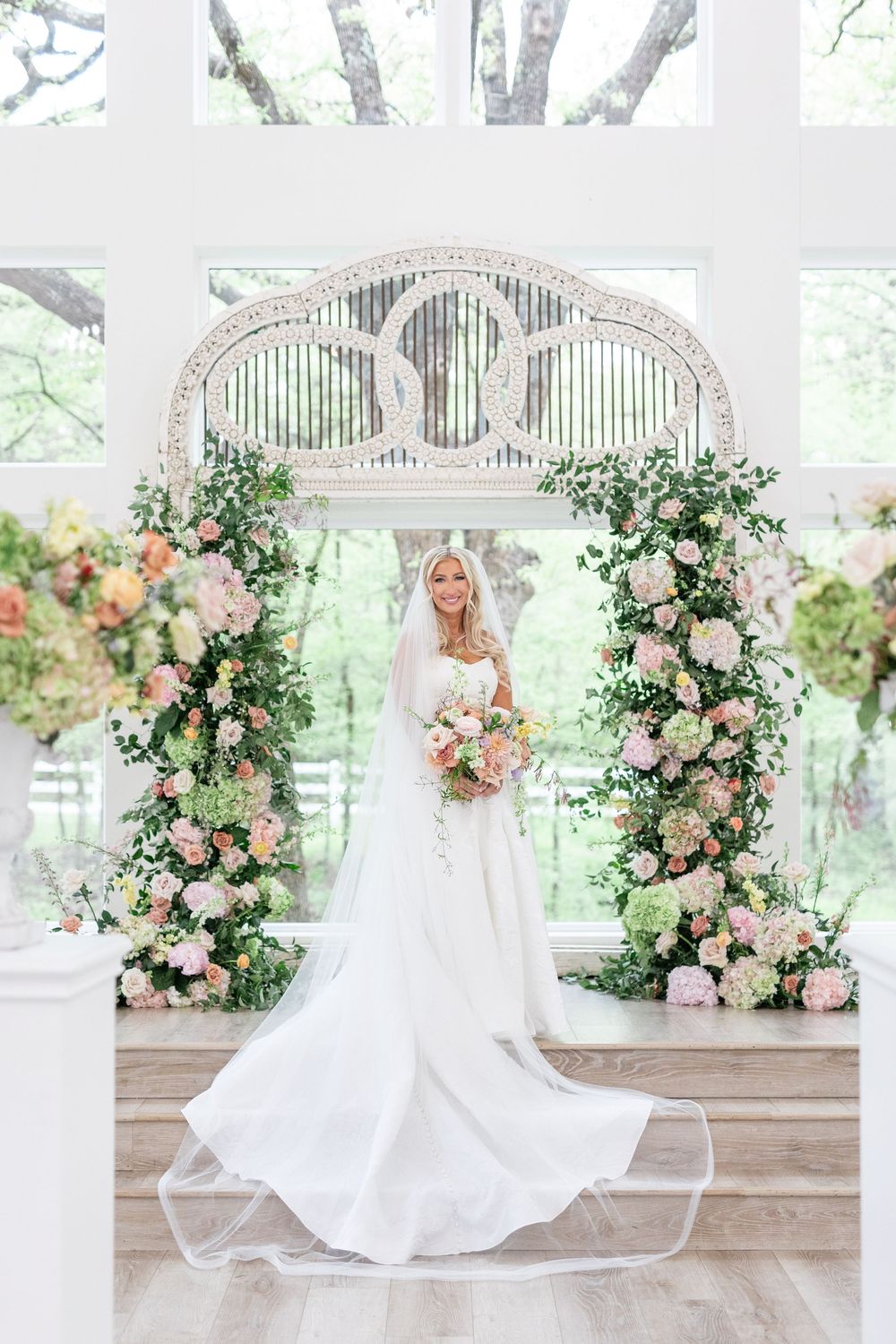 Smiling bride in white gown holding bouquet standing under floral arch with pink and peach blooms.