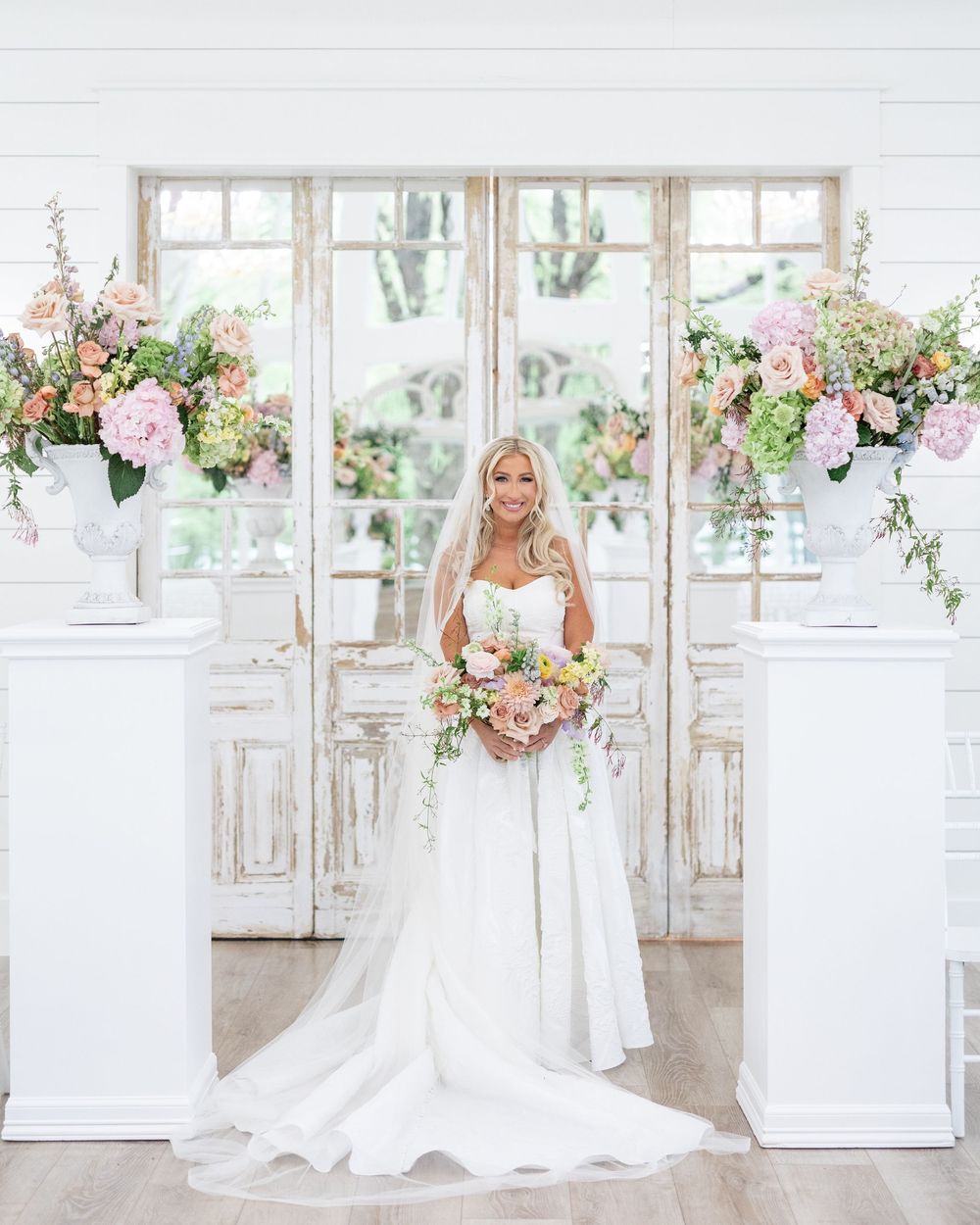 Elegant bride in strapless white gown holding pastel bouquet, standing before vintage doors with floral arrangements.