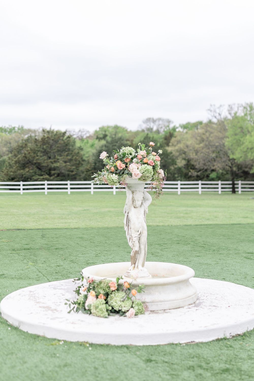 Classic cherub fountain statue adorned with peach roses and green hydrangeas on a manicured lawn.
