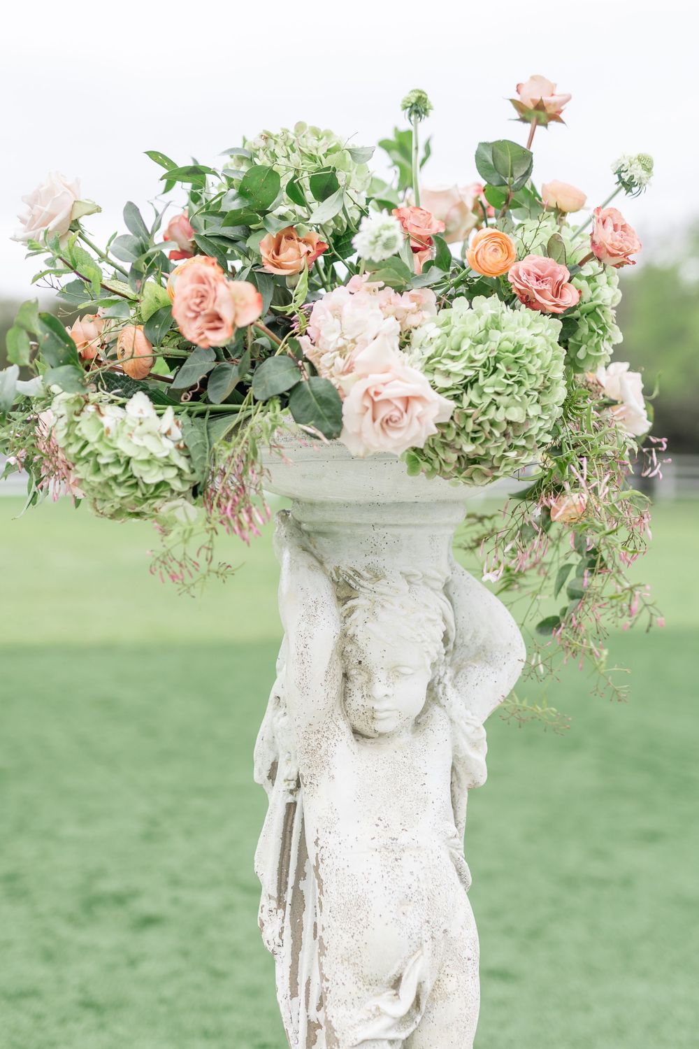 Stone cherub pedestal topped with lush arrangement of peach roses and green hydrangeas outdoors.