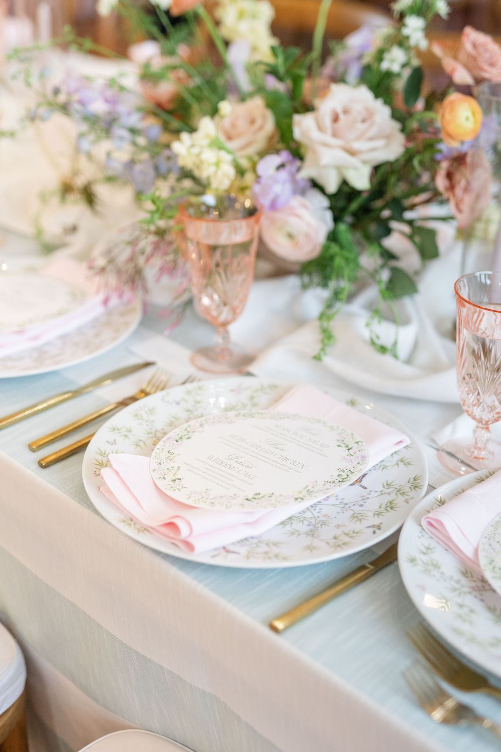 Elegant wedding place setting with floral china, pink napkin, gold flatware, and blush glassware.